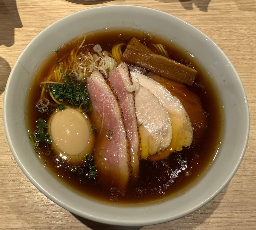 白い器に盛られた醤油ラーメン。澄んだ茶褐色のスープに、細めのストレート麺。トッピングは鴨チャーシューと鶏チャーシュー、味玉、メンマ、刻みネギ、青ネギ。上品で繊細な盛り付けが印象的。