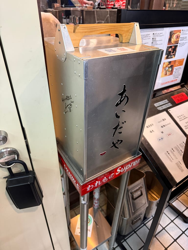 つけ麺専門店「あいだや」の店頭に置かれた岡持ち。店名のロゴが刻まれ、行列時の案内や演出に使われている。