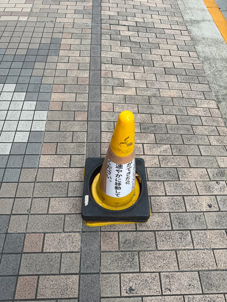 タイル張りの歩道に設置された黄色いカラーコーン。コーンには、行列時の通行マナーを呼びかける注意書きが貼られている。