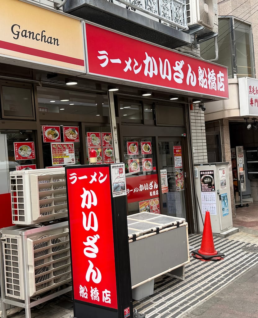 船橋駅近くにある「ラーメンかいざん 船橋店」の外観。赤い看板とメニュー写真が並ぶ店舗入口を夜に撮影した一枚。
